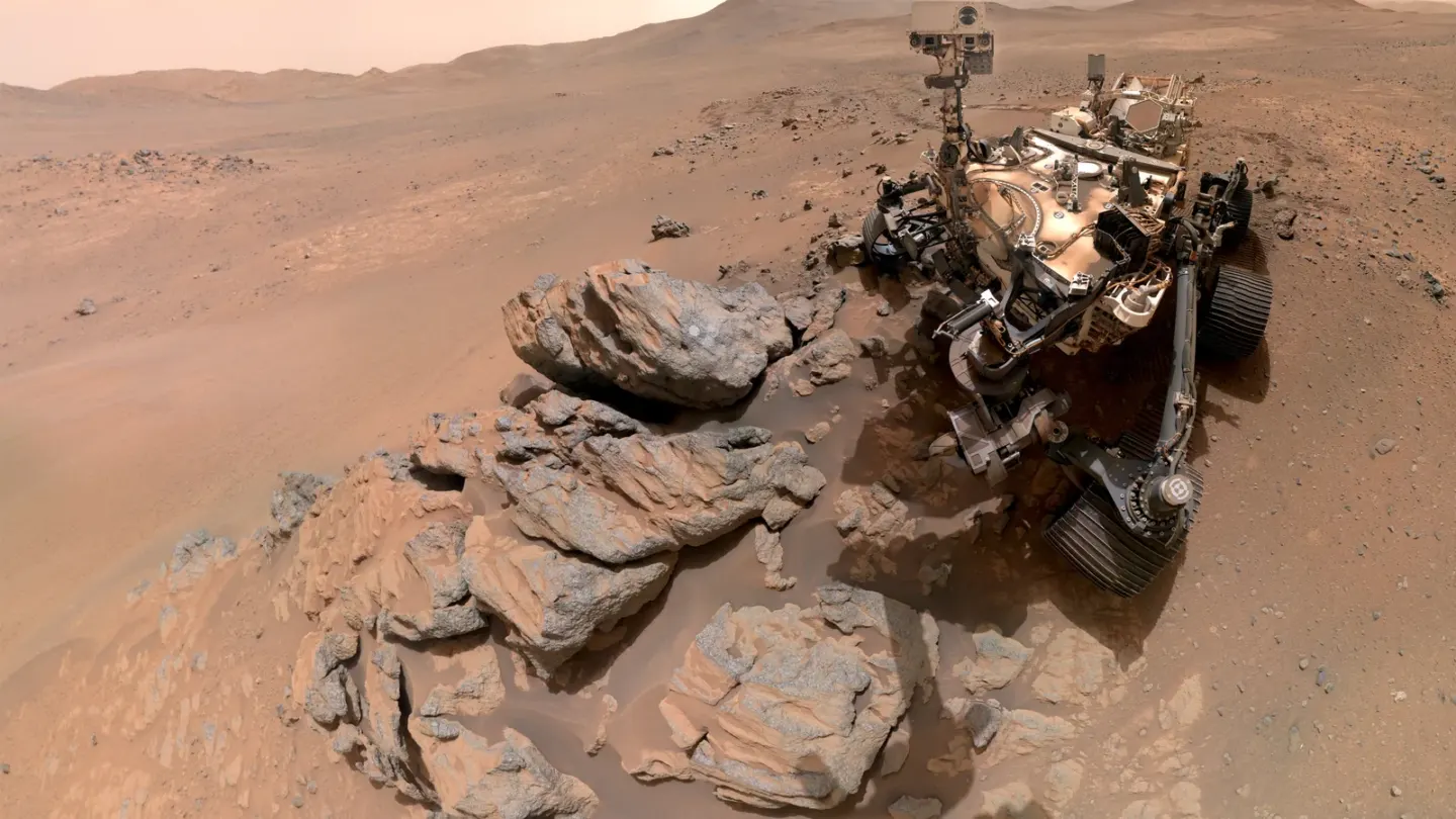 NASA-rover Perseverance tussen rotsen op Mars, met een uitgestrekt Marslandschap op de achtergrond.